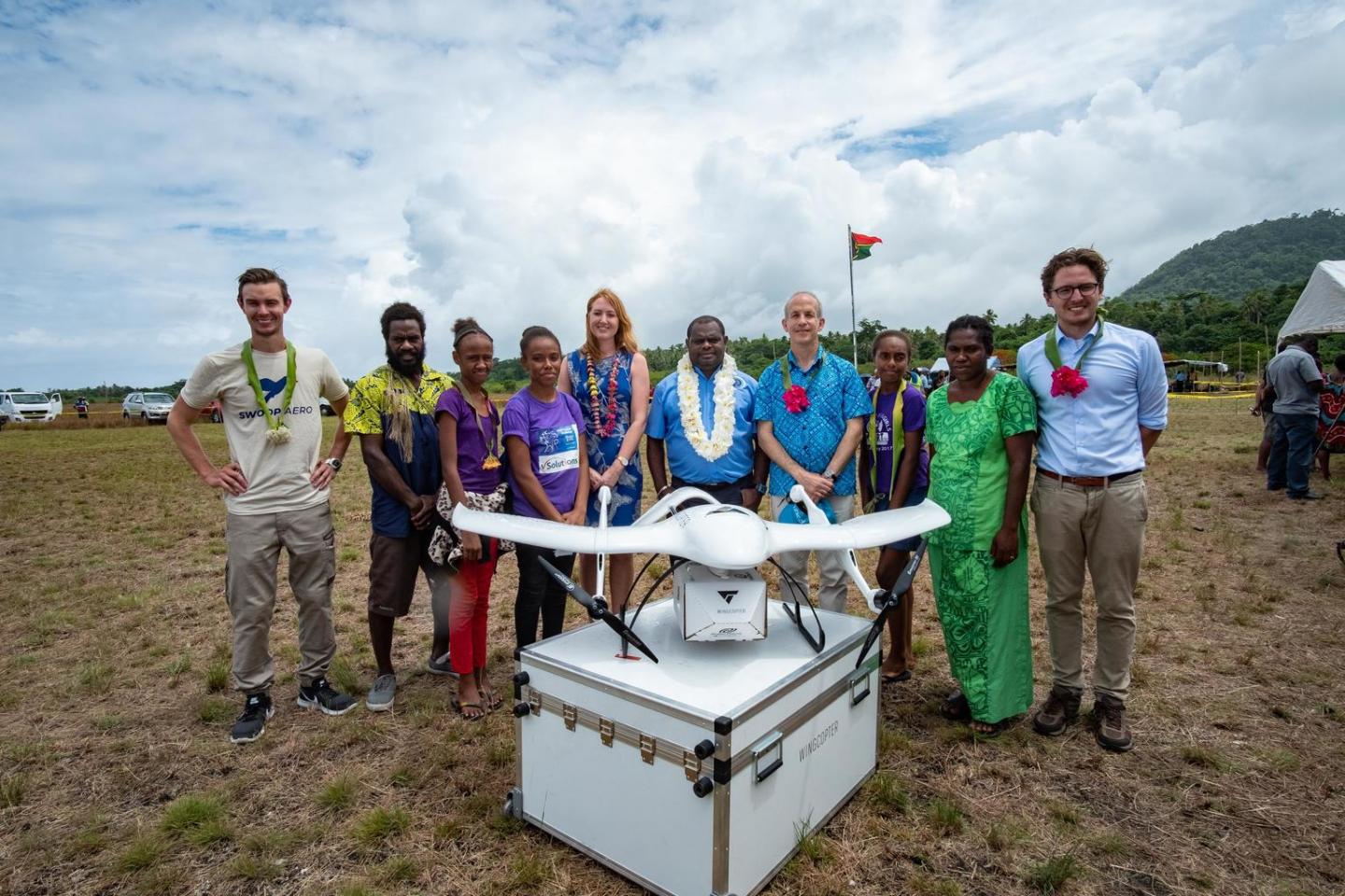 A group of ten people are standing in a field. A drone is placed in front of them on top of a crate.