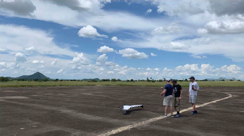 Three people stand on a large runway. The group is looking and pointing at a small drone on the runway.