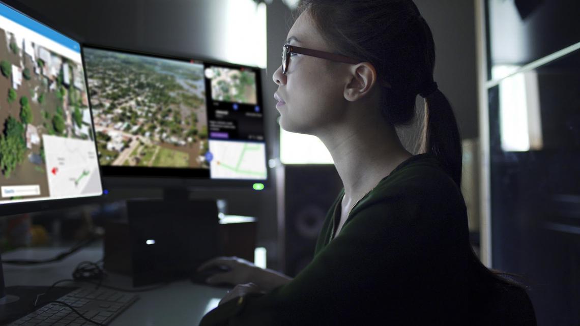 A woman is monitoring two computer screens. The screens show aerial imagery, perhaps taken from a drone.